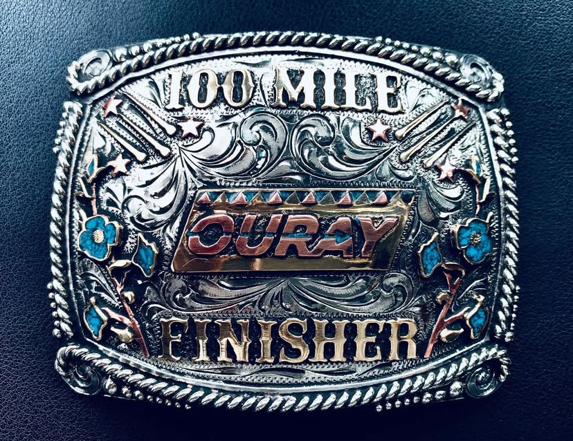 A large, ornate belt buckle with "100 Mile Finisher" and decorative engravings, as a prize given to runners who finish the Ouray 100 endurance run