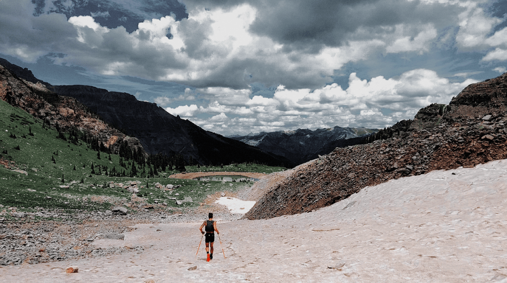 Ultra runner Alex Griffin in motion from afar descending down snowy mountainous slopes during the Ouray 100 ultramarathon with gray skies and dramatic scenery in the San Juan Mountains